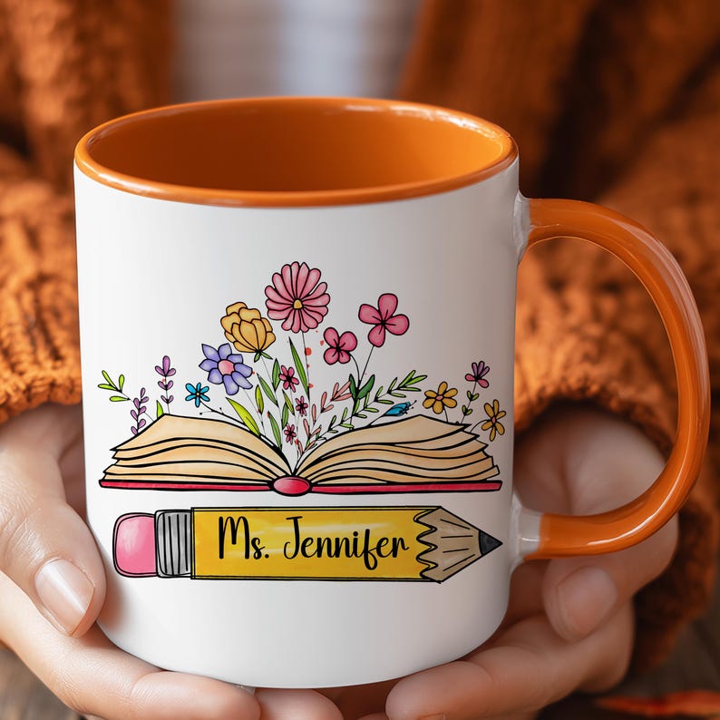 May include: A white ceramic mug with an orange interior and handle. The front features a graphic of an open book with colorful flowers blooming from the pages, positioned above a yellow pencil with the text Ms. Jennifer printed on it.