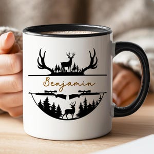 May include: White ceramic mug with a black rim and handle, featuring a deer and rifle design. The mug has the name "Benjamin" in a cursive font. The design includes silhouettes of trees and deer.