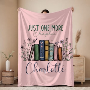 May include: A light pink throw blanket featuring the text "JUST ONE MORE Chapter" in dark green and black script. Below the text is an illustration of a row of colorful books surrounded by wildflowers. The name "Charlotte" is printed in large black script.