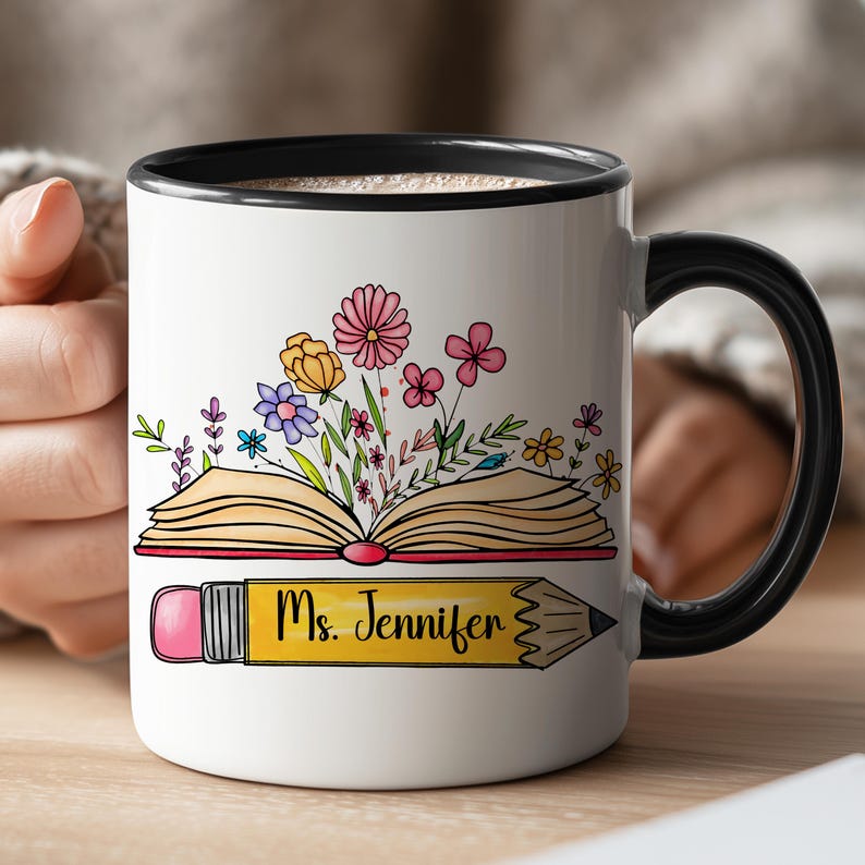 May include: A white ceramic mug with a black rim and handle, featuring a colorful illustration of an open book with blooming flowers emerging from the pages, positioned above a yellow pencil graphic with the text Ms. Jennifer printed on it.