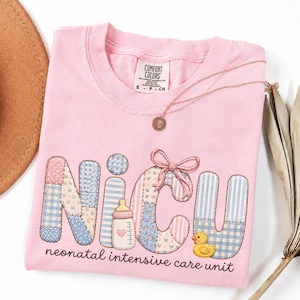 May include: A light pink t-shirt featuring the text NICU in a patchwork quilt style with blue, pink, and white patterns. The design includes a small baby bottle, a pink bow, and a yellow rubber duck. Below, the text reads neonatal intensive care unit.