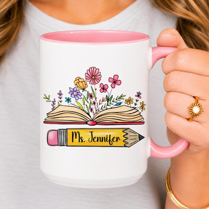 Personalized teacher mug featuring an open book with colorful blooming flowers and a custom name on a pencil. White ceramic mug with pink handle and interior, perfect teacher appreciation gift, back to school present, reading lover mug, and classroom coffee cup.