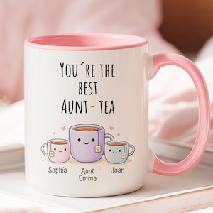 May include: White ceramic mug with a pink interior and handle. The mug features the text "YOU'RE THE BEST AUNT-TEA" and illustrations of three teacups with smiling faces. The names "Sophia", "Aunt Emma", and "Joan" are below the teacups.