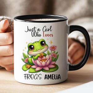 May include: White ceramic mug with a black handle and rim. The mug features a cartoon frog on a lily pad with the text "Just a Girl Who Loves FROGS AMELIA". The frog is green with large eyes and is surrounded by pink flowers.