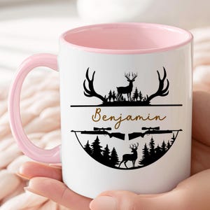 K&ouml;nnte beinhalten: Wei&szlig;e Keramiktasse mit rosa Innenraum und Henkel. Die Tasse zeigt ein schwarzes Hirschgeweih- und Wald-Design mit dem Namen "Benjamin" in goldener Schrift. Zwei Gewehre und ein weiteres Reh sind ebenfalls abgebildet.