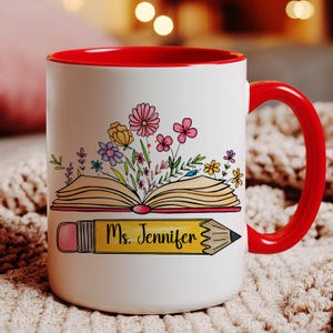 May include: A white ceramic mug with a bright red interior and handle. The front features a colorful illustration of an open book with various flowers blooming from the pages, positioned above a yellow pencil graphic with the text Ms. Jennifer.