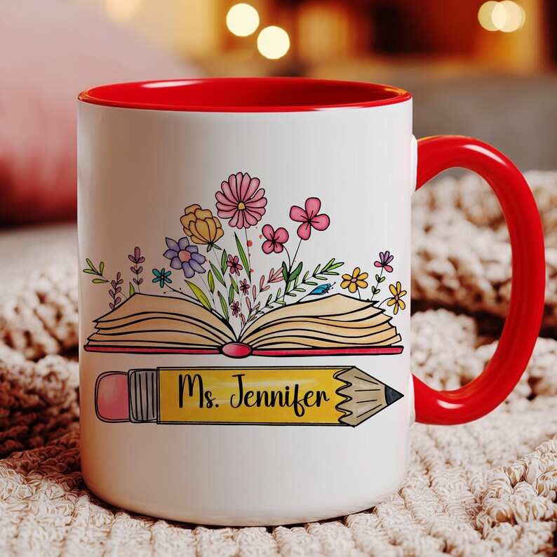 May include: A white ceramic mug with a bright red interior and handle. The front features a colorful illustration of an open book with various flowers blooming from the pages, positioned above a yellow pencil graphic with the text Ms. Jennifer.