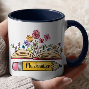 May include: A white ceramic mug with a dark blue interior and handle. The exterior features a graphic of an open book with colorful flowers emerging from the pages, positioned above a yellow pencil with the text Ms. Jennifer printed on it.