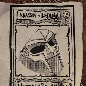 May include: A black and white illustration of a mask on a white card with the text "Mask of Doom" and "Armor: +2 to AC". The card has a black border and a small black and white illustration of a medal with the number "10" on it.