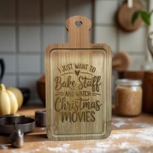 Op de afbeelding: Een rechthoekige bamboe snijplank met een handvat en de tekst "I JUST WANT TO Bake Stuff AND WATCH Christmas MOVIES" gegraveerd op het oppervlak. Het handvat heeft een hartvormige uitsparing. De plank is lichtbruin.