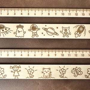 Op de afbeelding: Twee houten linialen met een schaalverdeling van 15 cm. De bovenste liniaal heeft een ruimtethema met een alien, een astronaut en een planeet. De onderste liniaal heeft een grillig ontwerp met een bloem, een meisje, een kat en een vlinder.