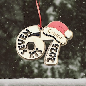 May include: Wooden Christmas ornament with the numbers "67" and "2023" cut out. The word "SEVEN SIX" is cut out within the number "67". A small Santa hat with the name "Conor" is attached. A red ribbon is at the top for hanging.