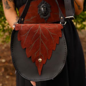 May include: Black leather crossbody bag with a rust-colored leaf-shaped flap. The leaf design has a darker brown vein pattern. The bag has a matching strap and a decorative belt. A brass-colored snap secures the flap.