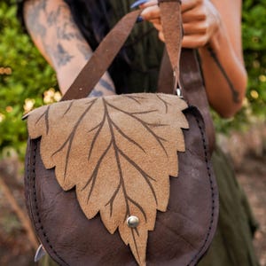 May include: A brown leather shoulder bag with a leaf design on the flap. The bag has a rounded shape and a strap. The leaf design is a lighter brown color and is stitched onto the darker brown leather. A silver button secures the flap.