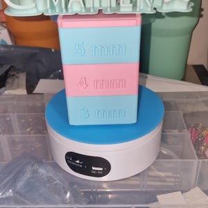 May include: A white rotating display stand with a blue base and a pink and blue stack of blocks with the measurements 5 mm, 4 mm, and 3 mm. The stand has a black label with the text "CW/OR/CCW" and "DC-5V". The stand is on a clear plastic storage box with compartments.