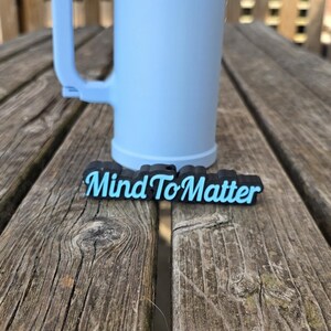 Könnte beinhalten: Ein hellblauer zylindrischer Becher mit einem stabilen Henkel und einem separaten schwarzen rechteckigen Anhänger, der den Text Mind To Matter in hellblauer Schrift zeigt. Der Becher hat eine glatte Oberfläche und einen flachen Boden.