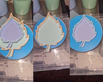 May include: A set of three leaf-shaped coasters. Each coaster features a layered design with a light blue base, a leaf outline in a light orange color, and a leaf interior in shades of green, purple, and light blue.