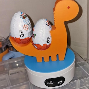 May include: A rotating display stand with a blue base and a white top. The stand features a bright orange dinosaur-shaped holder with two Kinder Surprise eggs inside. The stand has a power switch and a DC-5V port.