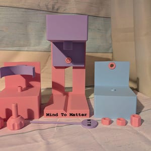May include: A collection of pastel-colored, geometric wooden shapes. The shapes include a tall, pink and purple structure, a pink chair, a blue chair, and various pink nuts and bolts. The text "Mind To Matter" is visible on a purple tag.