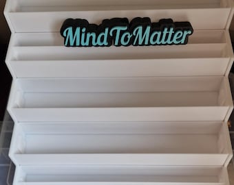 May include: A white, tiered display shelf with multiple levels. The shelf has a rectangular shape and is made of a smooth, solid material. The text "Mind To Matter" is displayed in black and teal on the top shelf.