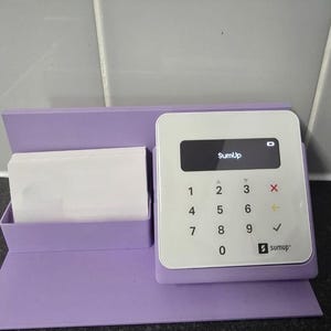 May include: A white SumUp card reader with a black screen displaying "SumUp" and a keypad. It sits in a lilac holder with a compartment for white cards. The SumUp logo is visible.