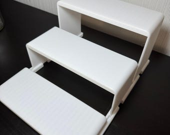 May include: A white, three-tiered display stand. The stand has a stepped design, with each level slightly smaller than the one below. The surfaces are smooth and the edges are rounded. The stand is designed to elevate items for display.