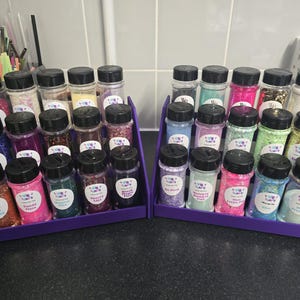 May include: Two purple tiered display stands filled with small clear jars of glitter with black lids. The jars contain glitter in various colors, including pink, blue, green, and gold. Each jar has a white label with text.