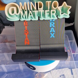 May include: Two 3D printed phone stands with the words "@MIND TO MATTER" and a star on top. The stands are grey with black bases and have the names "RISTAN" and "BRAX" printed on them in orange and blue.