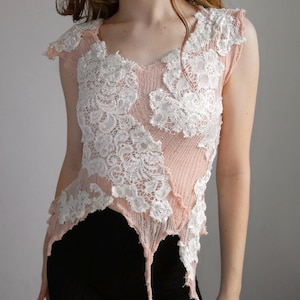 May include: A sleeveless top in a soft peach color featuring a textured, ribbed knit fabric. The garment is adorned with asymmetrical white floral lace appliqués stitched across the front, creating a layered look with a jagged, uneven hemline.
