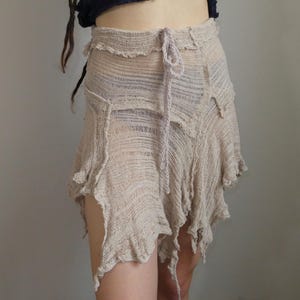 May include: Beige, asymmetrical skirt with a frayed hemline. The skirt has a drawstring waist and a layered, textured appearance. The fabric appears to be a loosely woven material, creating a unique, bohemian style.