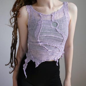 May include: A sleeveless lavender top made of a sheer, open-knit fabric with a distressed, raw-edge construction. The garment features visible seams, an asymmetrical hemline, and a circular cutout detail on the upper chest area.