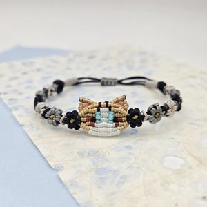 May include: A black string bracelet with a woven cat face charm and small flower accents. The cat face is brown, white, and blue.