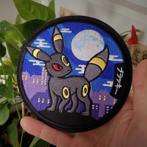Könnte beinhalten: Runder, gestickter Aufnäher mit einem schwarzen Umbreon-Charakter mit gelben Akzenten, roten Augen und einer Stadtlandschaft vor einem Vollmond. Der Aufnäher hat japanischen Text und einen schwarzen Rand.