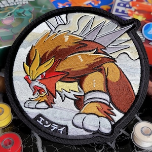 Peut inclure: Écusson brodé représentant Entei, un Pokémon de feu, dans des tons de marron, de rouge et de jaune, sur un fond blanc et gris. L'écusson a une bordure noire et du texte japonais. L'écusson est rond et est probablement destiné à une veste ou un sac.