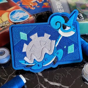 May include: A rectangular embroidered patch featuring a blue creature with a light gray shell and a long neck. The design includes geometric blue background patterns and small teal diamond accents, with a dark blue border surrounding the shape.