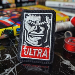 Può includere: Toppa ricamata in bianco e nero con un viso bianco e la parola "ULTRA" in lettere rosse.