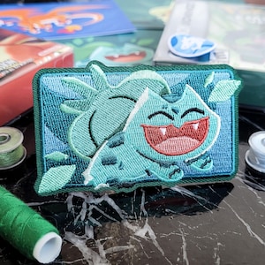 May include: Embroidered patch with a Bulbasaur design in teal, green, and red. The rectangular patch has a teal border and shows the Pokémon character smiling. Sewing supplies and game boxes are in the background, with thread spools visible.