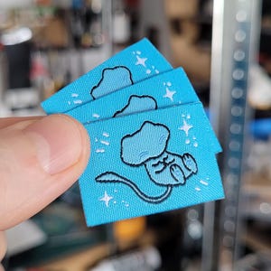 Peut inclure: Trois petites étiquettes en tissu bleu clair avec une illustration de créature de dessin animé. La créature est bleu clair avec un contour noir, et les étiquettes ont des étoiles blanches. Les étiquettes sont tenues dans une main.