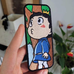Peut inclure: Un écusson brodé rectangulaire représentant un personnage de dessin animé aux cheveux noirs avec une petite couronne jaune, portant une tunique bleue et des leggings blancs. Le personnage est devant des troncs d'arbres et des feuilles vertes.