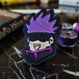 May include: Purple and black embroidered patch of a cartoon character with a purple mohawk, black eyes, and a white face. The character is smiling and has a pink mouth.