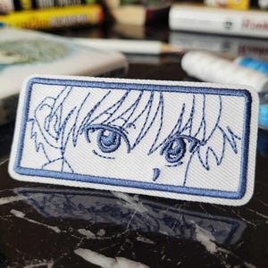 Könnte beinhalten: Gestickter Aufnäher mit Augen und Haaren eines Anime-Charakters in Blau und Weiß. Der rechteckige Aufnäher hat einen blauen Rand, geeignet für Kleidung oder Accessoires. Das Design ist detailliert, mit sichtbaren Nähten.