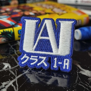 Può includere: Una toppa ricamata blu e bianca con la lettera "A" all'interno di un quadrato e il testo "クラス 1-A" sotto.