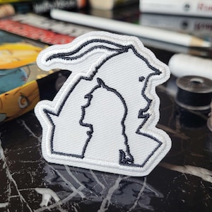Peut inclure: Écusson brodé blanc représentant deux silhouettes avec des contours noirs. Le patch a une bordure noire et est probablement thermocollant ou à coudre. Le patch est sur une surface en marbre noir et blanc.