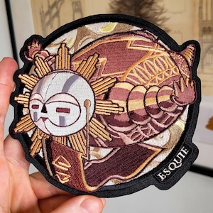 Peut inclure: Écusson brodé avec un motif de visage de soleil dans des tons de rouge et d'or. Le patch a une bordure noire et le mot "ESQUIE" est brodé en bas à droite. Le patch est tenu dans une main.