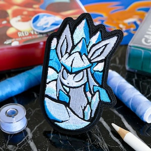 May include: An embroidered patch featuring a stylized Glaceon character in shades of light blue, white, and dark gray. The design is enclosed within a thick black border, showcasing intricate stitching that defines the character's ears and body.