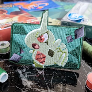 Peut inclure: Un écusson brodé rectangulaire représentant une créature vert clair aux yeux rouges avec un motif en losange rouge sur la poitrine. Le fond de l'écusson présente un motif géométrique vert foncé avec de petits accents violets et une bordure vert foncé.