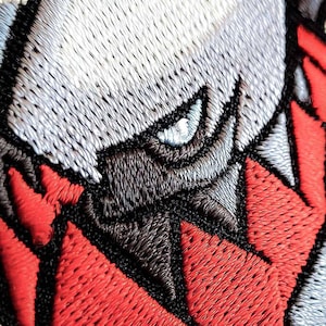 Op de afbeelding: Close-up van een geborduurde patch met een gestileerd oog en gezichtskenmerken. Het ontwerp gebruikt grijstinten, zwart en rood, met gedetailleerde steken. De patch heeft een getextureerde uitstraling.