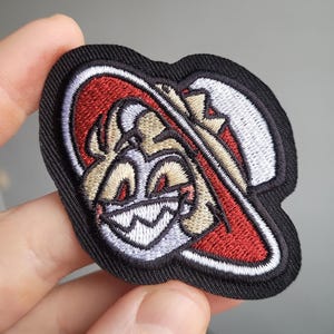 Peut inclure: Écusson brodé représentant un personnage de dessin animé souriant avec des yeux rouges et un chapeau. Le patch a une bordure noire et est principalement rouge, blanc et beige. Le design est détaillé et semble être brodé à la machine.