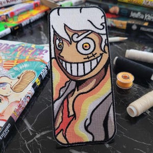 May include: Embroidered patch featuring a character with white hair, a mischievous grin, and a colorful background. The character's eye is visible, and the patch is detailed with intricate stitching.