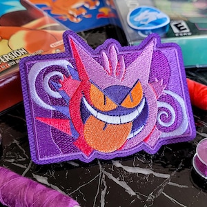 Peut inclure: Un écusson rectangulaire brodé représentant le personnage Gengar violet avec des yeux orange et un large sourire, montrant une longue langue orange. Le motif inclut des accents rouges et violet clair, avec des spirales sur fond violet.
